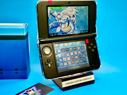A fully modded blue/black Nintendo 3DS XL with custom firmware shows a Blastoise wallpaper on the top screen, app icons below, and stands on a CANADA CUSTOM CONSOLES stand with a blue box and business card nearby.