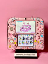 A Cute Pink Nintendo 2DS — Japanese Edition | Fully Modded, decorated with stickers, rests on a CANADA CUSTOM CONSOLES stand against a pink background. The top screen displays a pastel animal theme, suggesting pre-installed games.