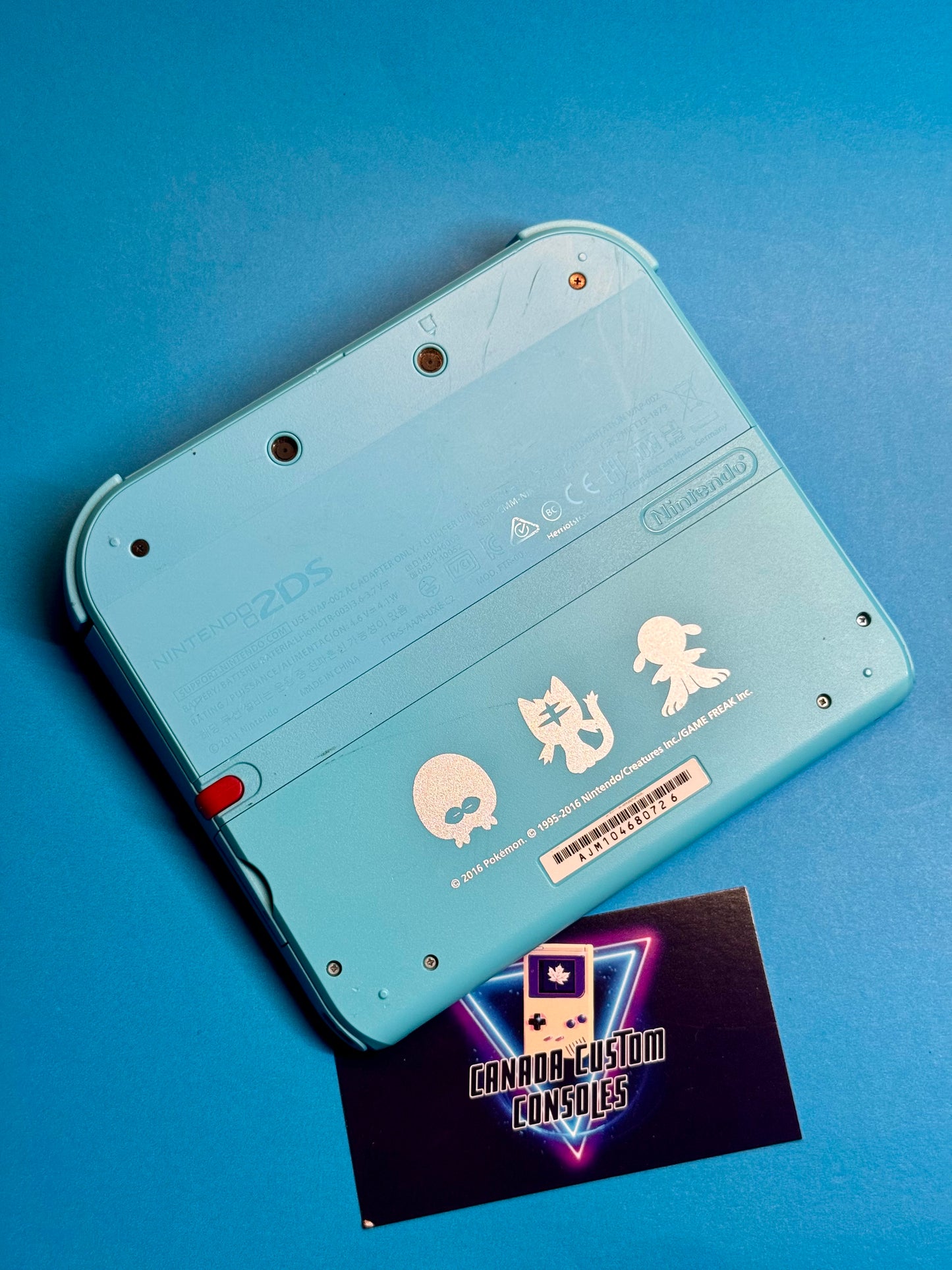 A Nintendo 2DS — Pokémon Sun & Moon Edition in turquoise with three white character decals on the back rests on a blue surface next to a Canada Custom Consoles business card.