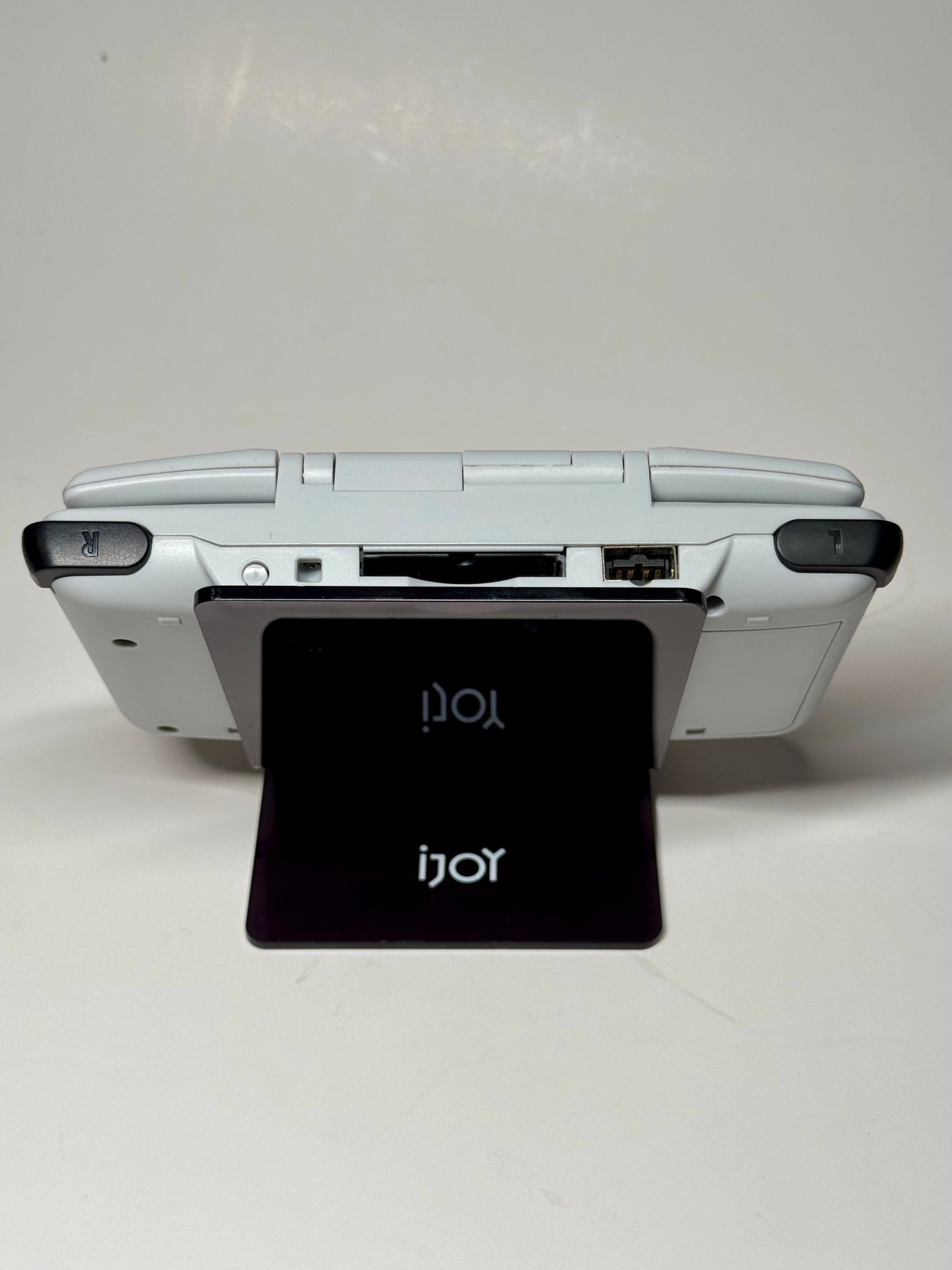 The Nintendo DS – SNES Edition, a white handheld in an SNES-style shell, is displayed on a black iJoy card stand. Viewed from the back, its cartridge slot and shoulder buttons evoke retro gaming nostalgia.