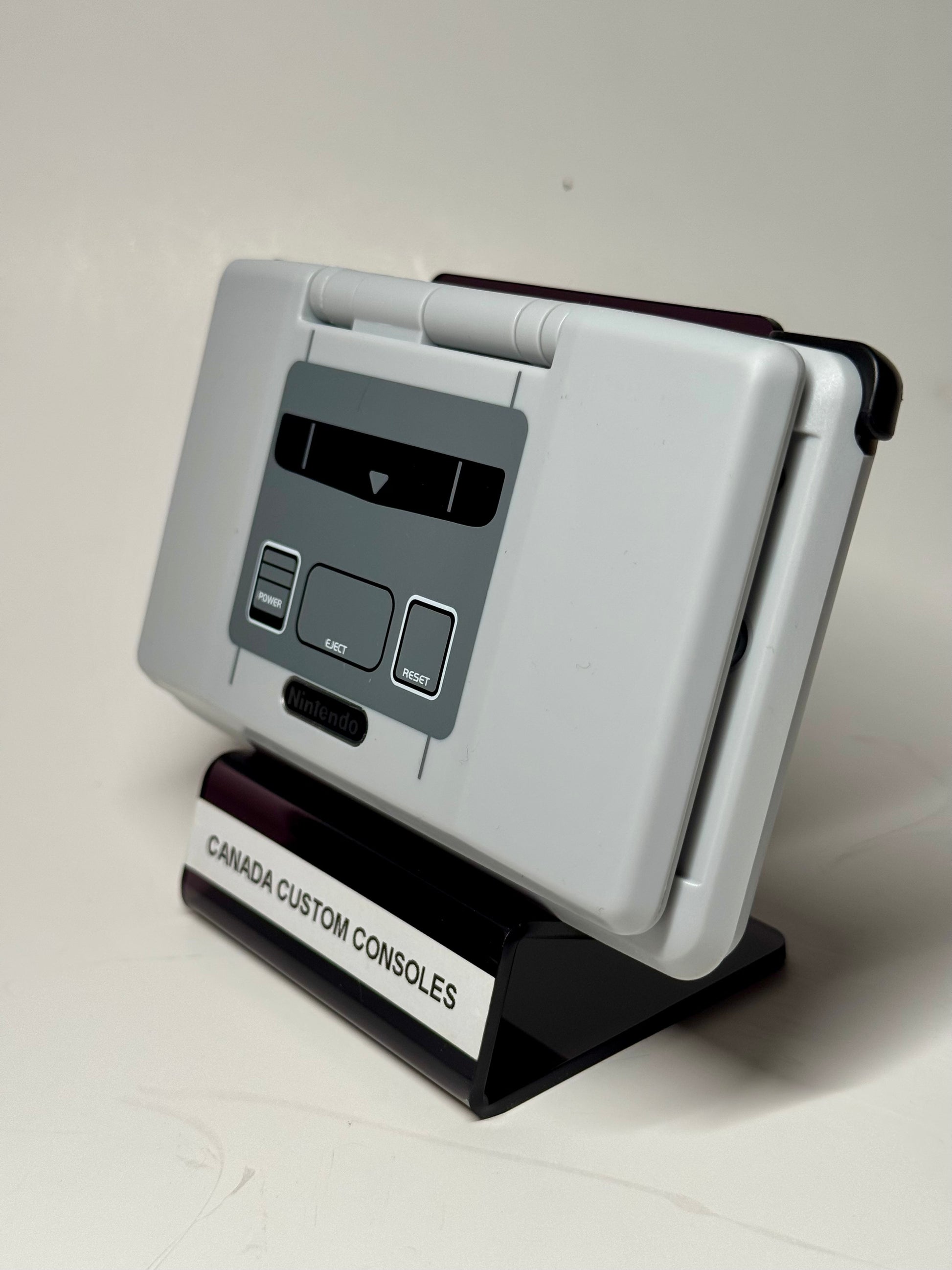 The Nintendo DS – SNES Edition, with a retro-inspired shell, is showcased on a stand labeled CANADA CUSTOM CONSOLES against a plain white background.