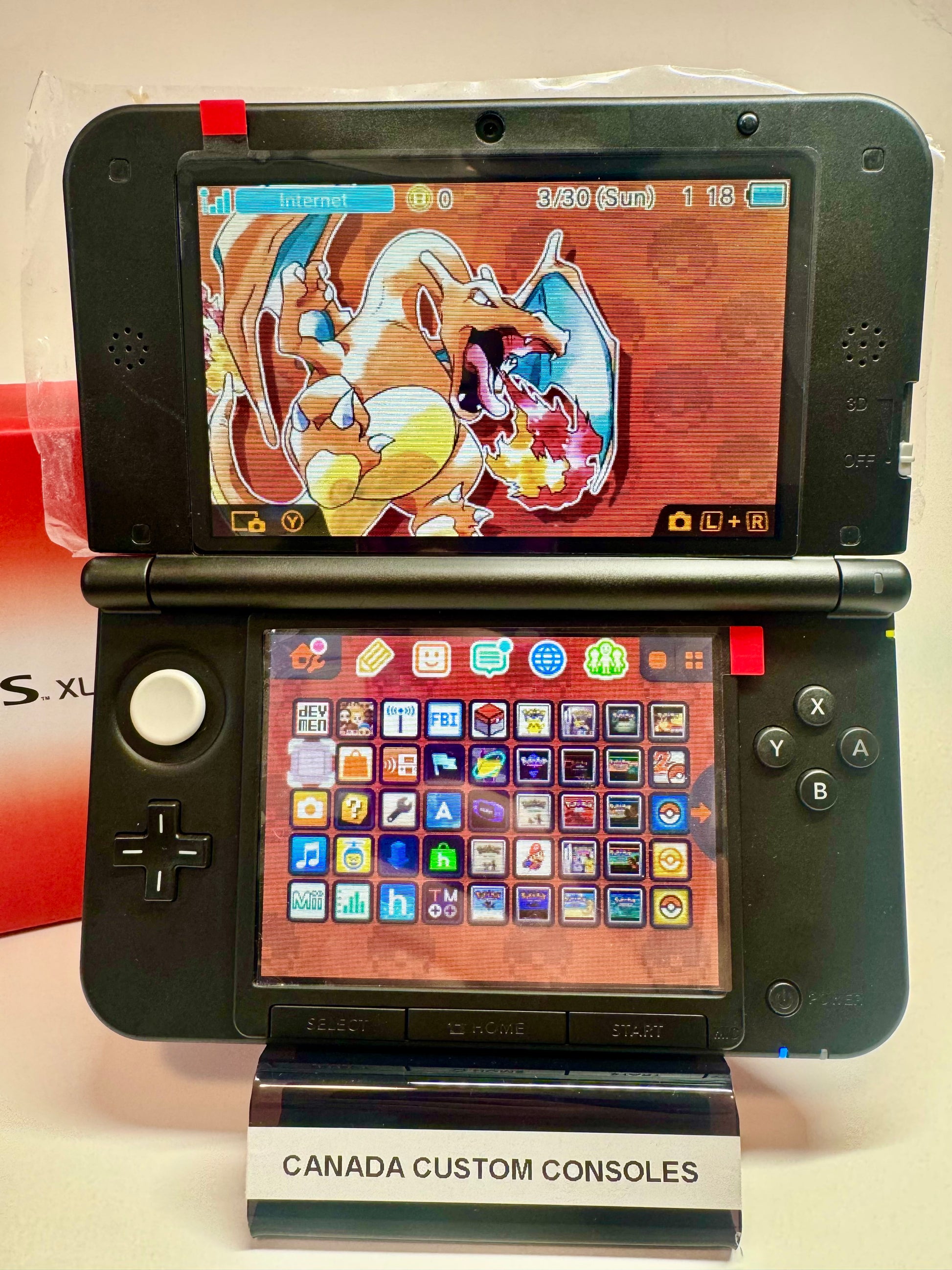 A red/black refurbished Nintendo 3DS XL—fully modded—features a Charizard-themed top screen, a colorful app icon grid, and sits on a stand labeled CANADA CUSTOM CONSOLES.
