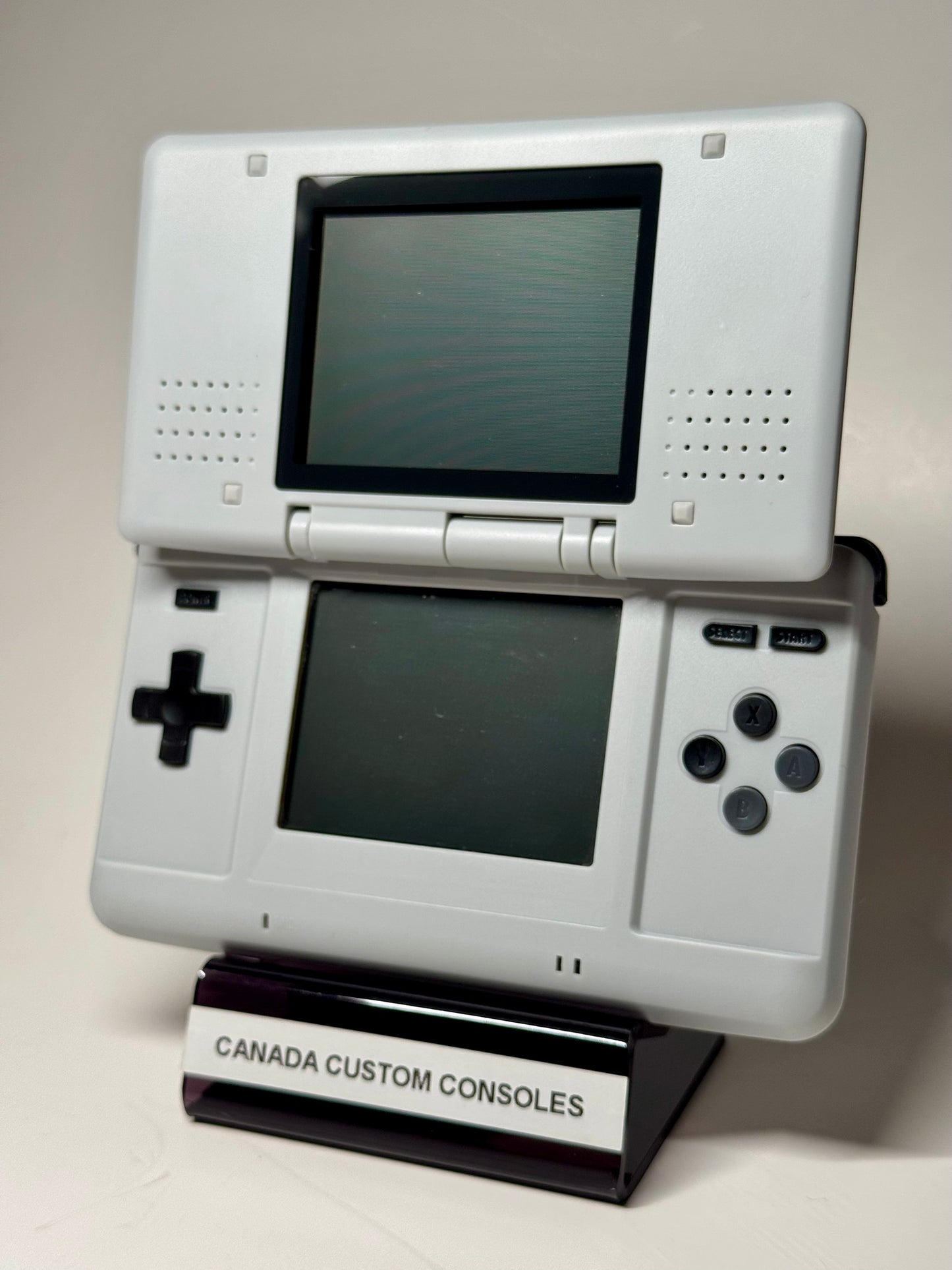 The Nintendo DS – SNES Edition, a white handheld with dual screens, stands upright on a CANADA CUSTOM CONSOLES stand against a plain background—ideal for retro gaming fans.