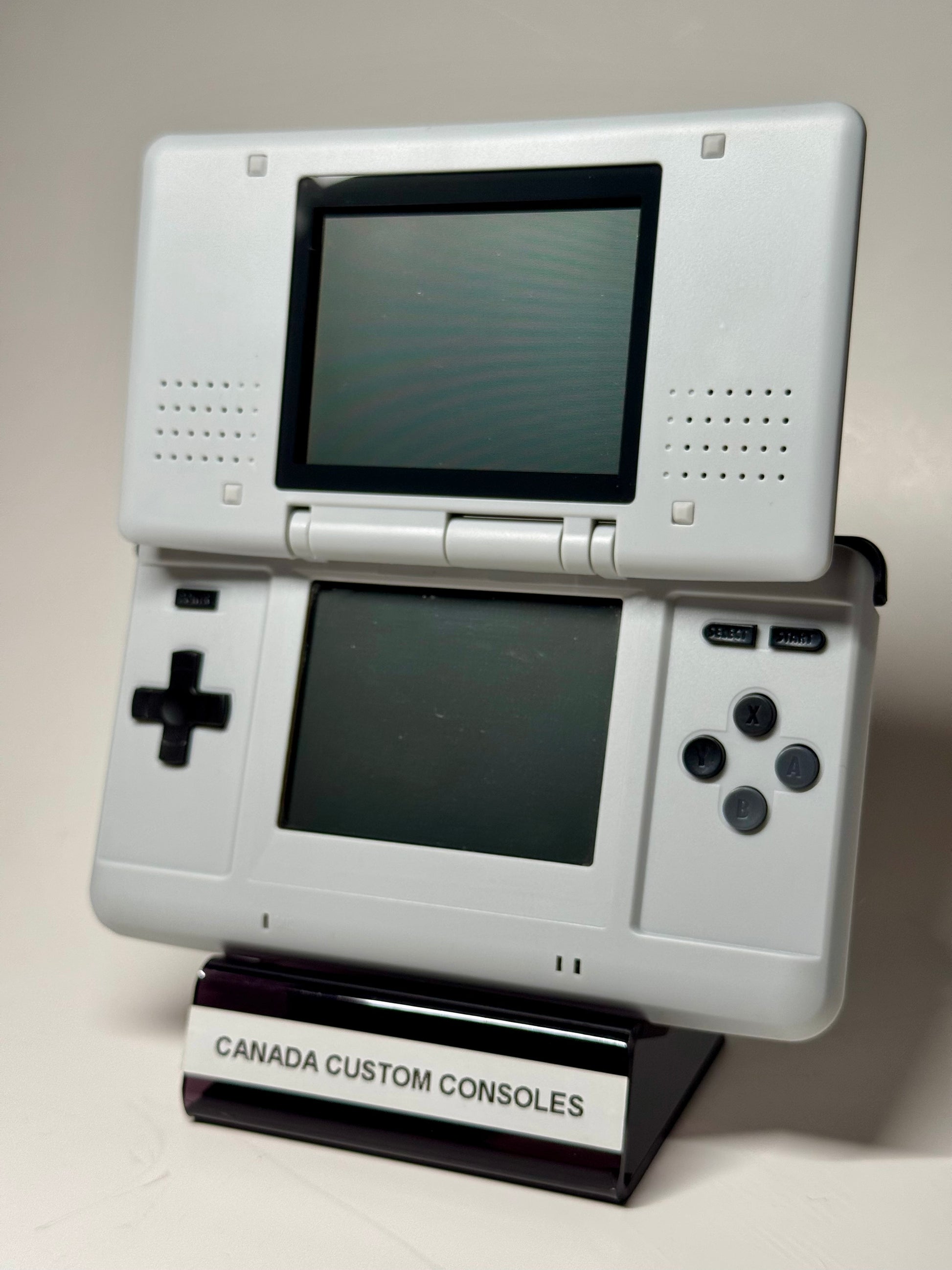 The Nintendo DS – SNES Edition, a white handheld with dual screens, stands upright on a CANADA CUSTOM CONSOLES stand against a plain background—ideal for retro gaming fans.
