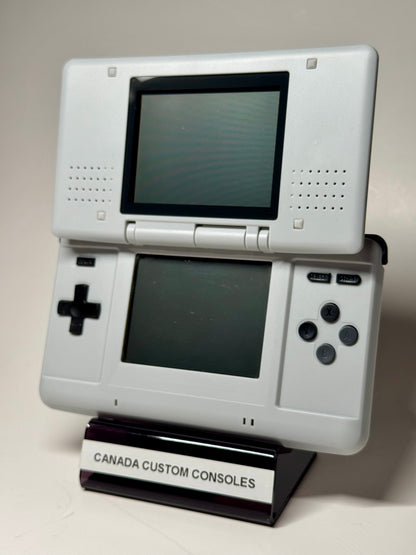 The Nintendo DS – SNES Edition, a white handheld with dual screens, stands upright on a CANADA CUSTOM CONSOLES stand against a plain background—ideal for retro gaming fans.