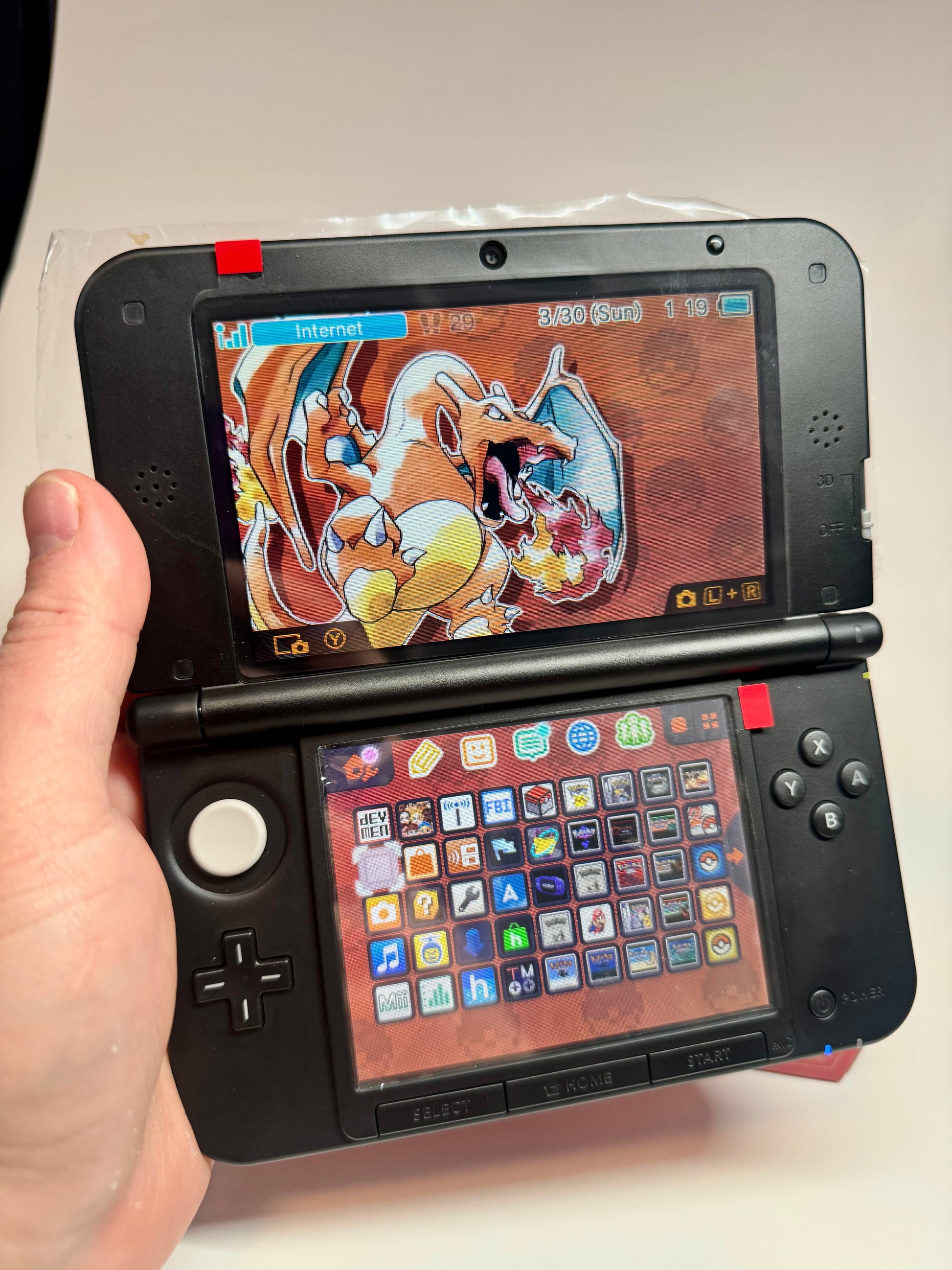 A hand holds an open refurbished Nintendo 3DS XL — Red/Black (fully modded), showcasing a Charizard-themed top screen and app icons below, with the date and time displayed on this stylish retro gaming console.