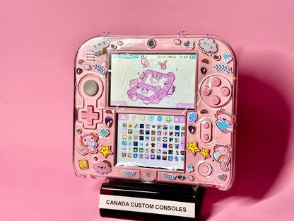 A Cute Pink Nintendo 2DS — Japanese Edition | Fully Modded, adorned with colorful cloud, star, and animal stickers, sits on a stand labeled CANADA CUSTOM CONSOLES against a solid pink background.