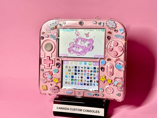A Cute Pink Nintendo 2DS — Japanese Edition | Fully Modded, adorned with colorful cloud, star, and animal stickers, sits on a stand labeled CANADA CUSTOM CONSOLES against a solid pink background.