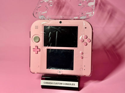 A Cute Pink Nintendo 2DS — Japanese Edition | Fully Modded is shown with a protective cover on a black CANADA CUSTOM CONSOLES stand, set against a solid pink background.