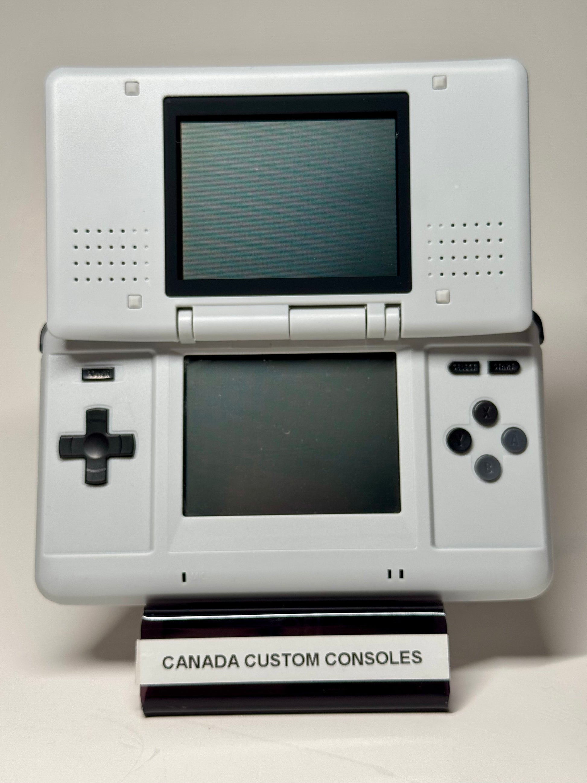 The Nintendo DS – SNES Edition is a white handheld console with dual screens, control buttons in an SNES-style shell, and a stand labeled CANADA CUSTOM CONSOLES, shown on a plain background.