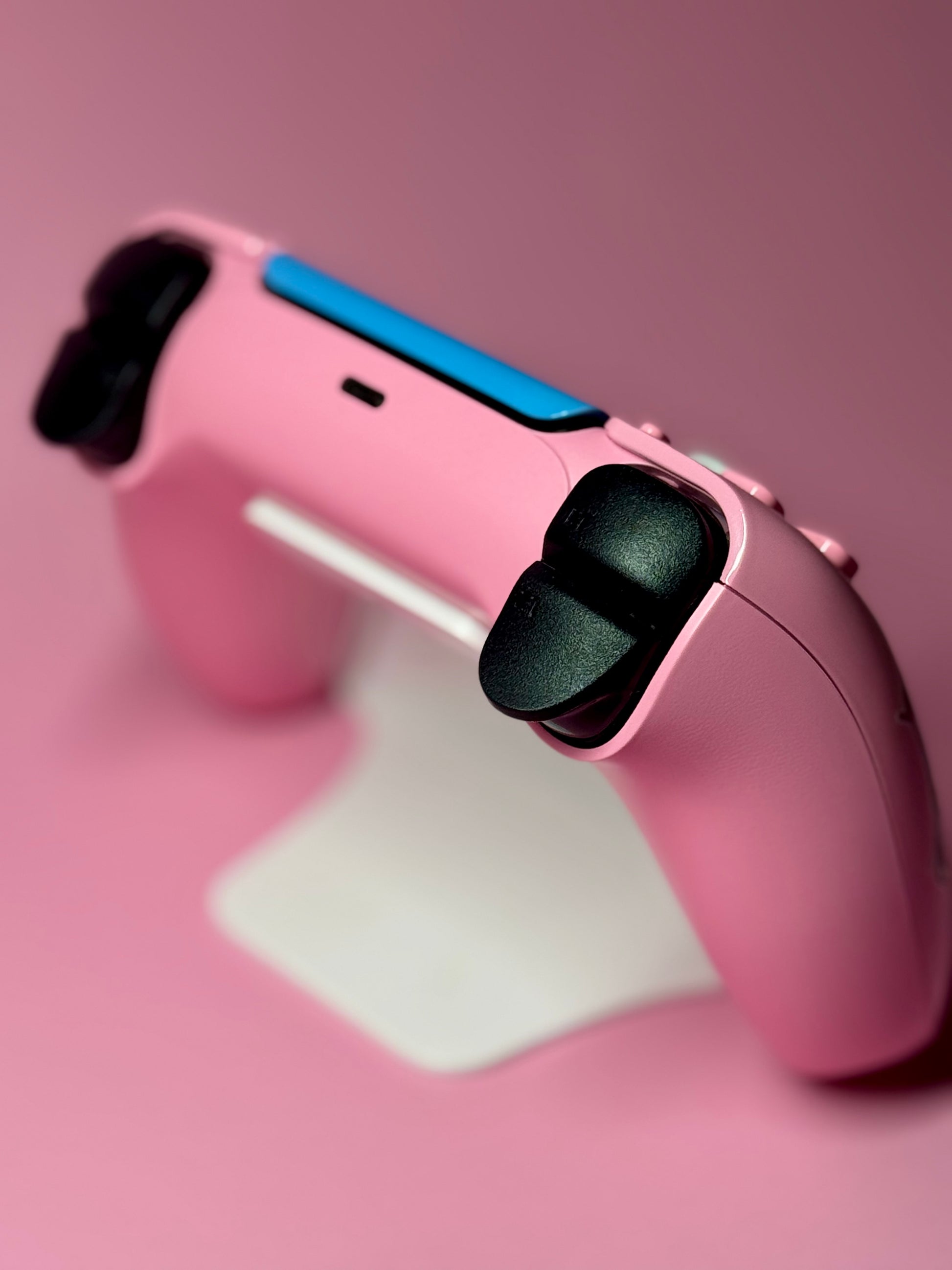 A D.Va Overwatch Custom PS5 Controller with Hall Effect sticks, finished in pink with blue accents, rests on a white stand against a pink background.