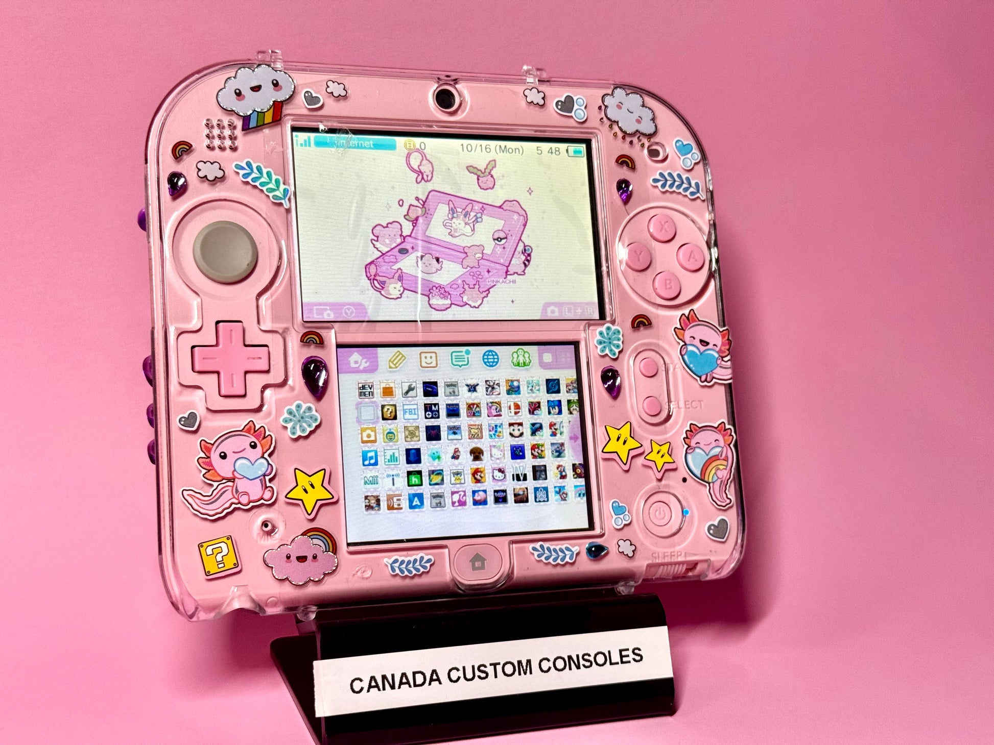 A Cute Pink Nintendo 2DS — Japanese Edition | Fully Modded sits on a stand labeled CANADA CUSTOM CONSOLES, decorated with unicorn, rainbow, heart, and star stickers against a matching pink background and showing an adorable pastel theme.