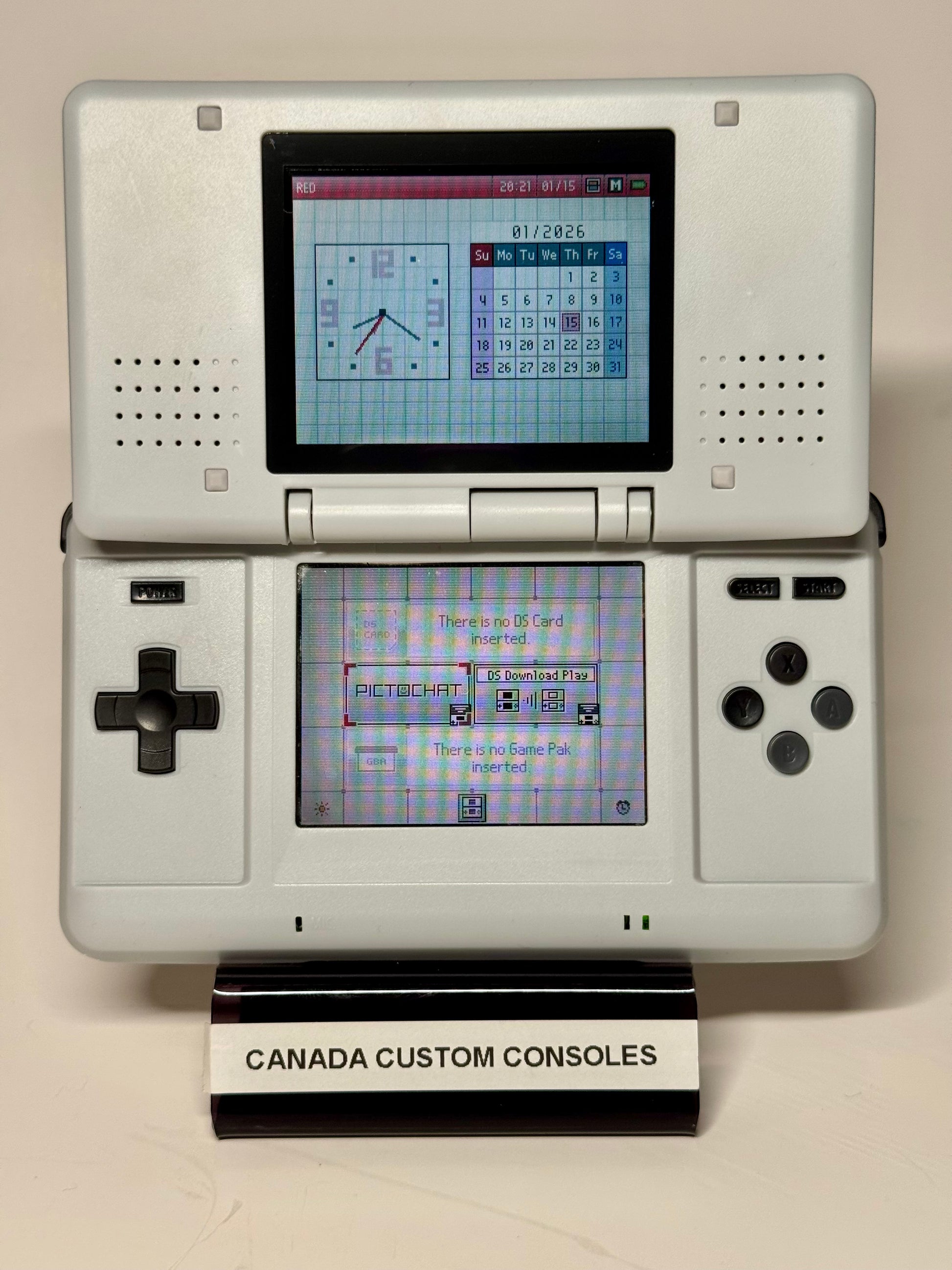An open, powered-on Nintendo DS – SNES Edition displays clock and calendar screens on a black-and-white stand labeled CANADA CUSTOM CONSOLES—a must-have for retro gaming fans.