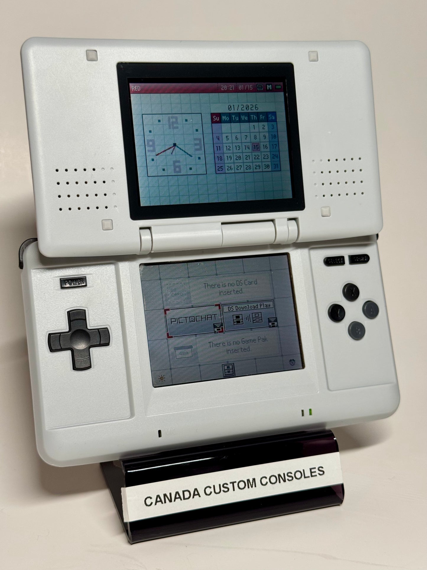 The Nintendo DS – SNES Edition, featuring a SNES-inspired white shell, is displayed on a black CANADA CUSTOM CONSOLES stand with its screens showing the menu and calendar—a standout piece for any retro gaming collection.