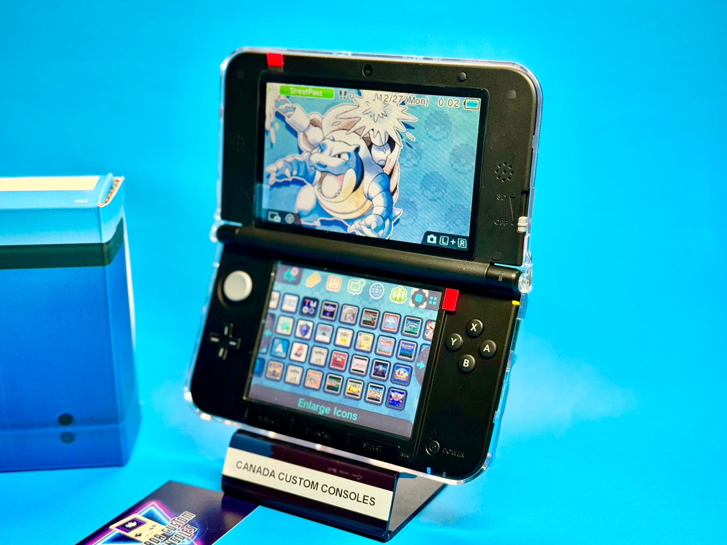 A fully modded Nintendo 3DS XL — Blue/Black is on display with a Pokémon-themed screen, standing on a clear Canada Custom Consoles stand against a bright blue background.