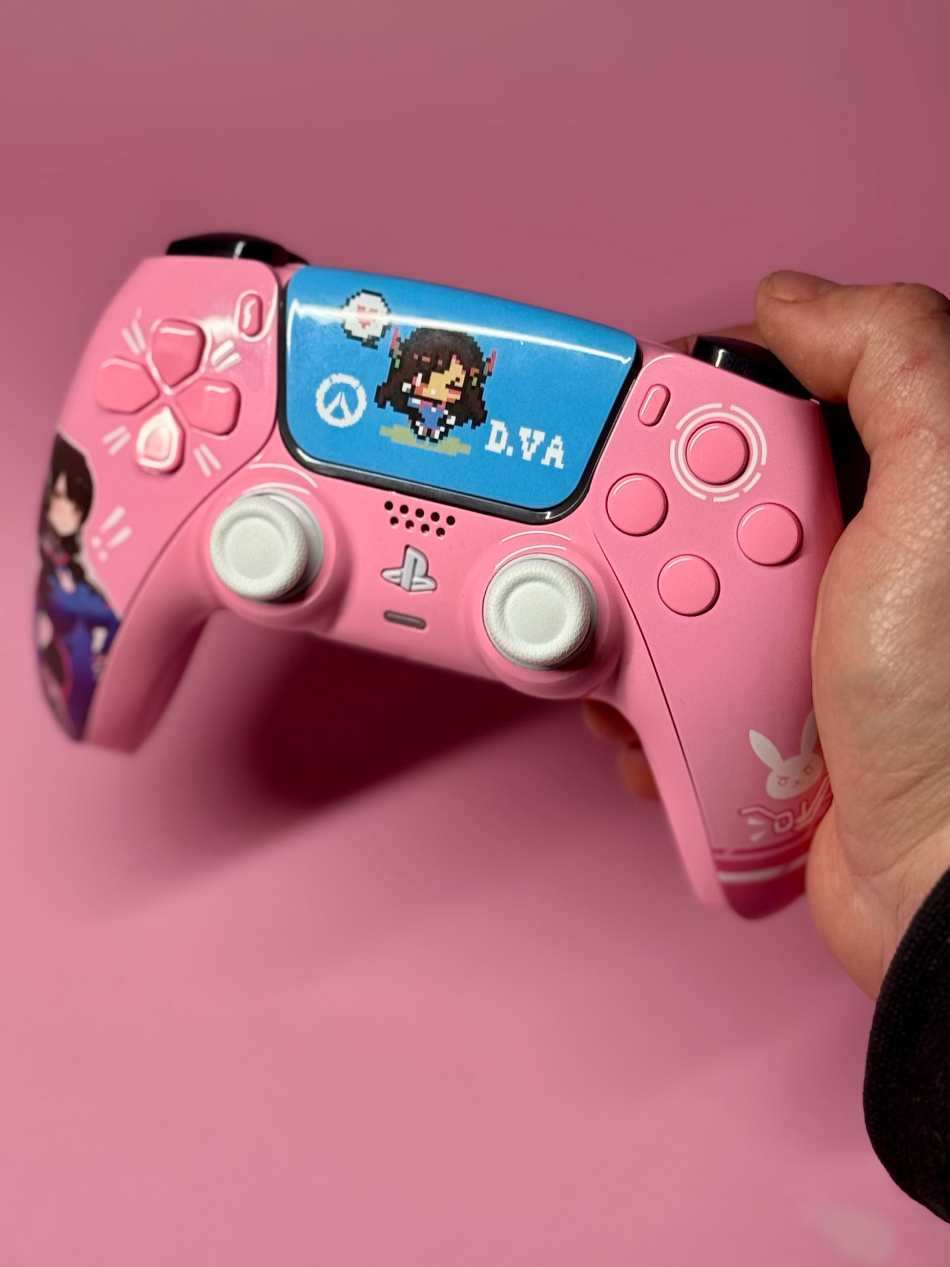 A hand holds the D.Va Overwatch Custom PS5 Controller | Hall Effect Sticks | Pink, featuring pixel art of D.Va on the touchpad and themed decals, all set against a pink background for the ultimate Overwatch look.