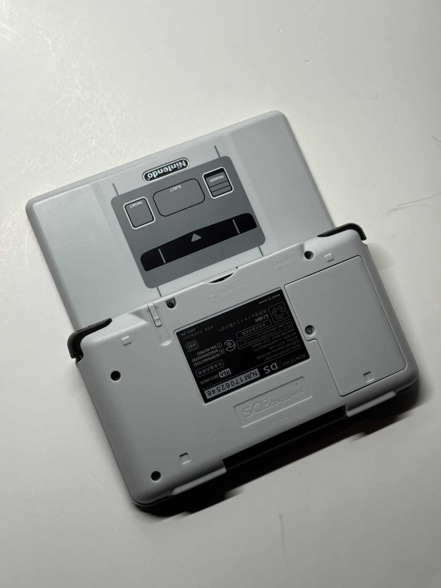 A close-up photo of a gray Nintendo DS – SNES Edition handheld console from the back, showing the cartridge slot and labels, partially open on a white surface—ideal for retro gaming fans.