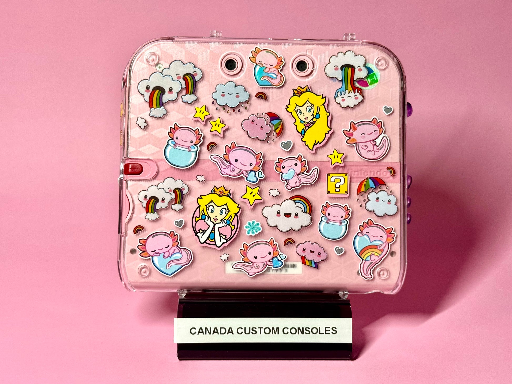 The Cute Pink Nintendo 2DS — Japanese Edition | Fully Modded features colorful stickers of clouds, rainbows, stars, axolotls, and a princess. Displayed on a CANADA CUSTOM CONSOLES stand with a pink backdrop, it includes region-free custom firmware.