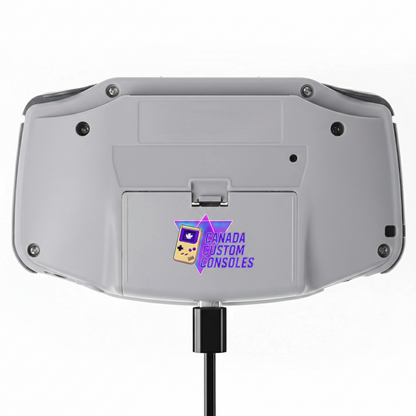 The image shows the back of a Game Boy Advance – Wario Edition (IPS 3.0 Laminated Display, Custom Build) with a Canada Custom Consoles logo—maple leaf and retro controller—by the battery compartment and a cable plugged into the bottom.